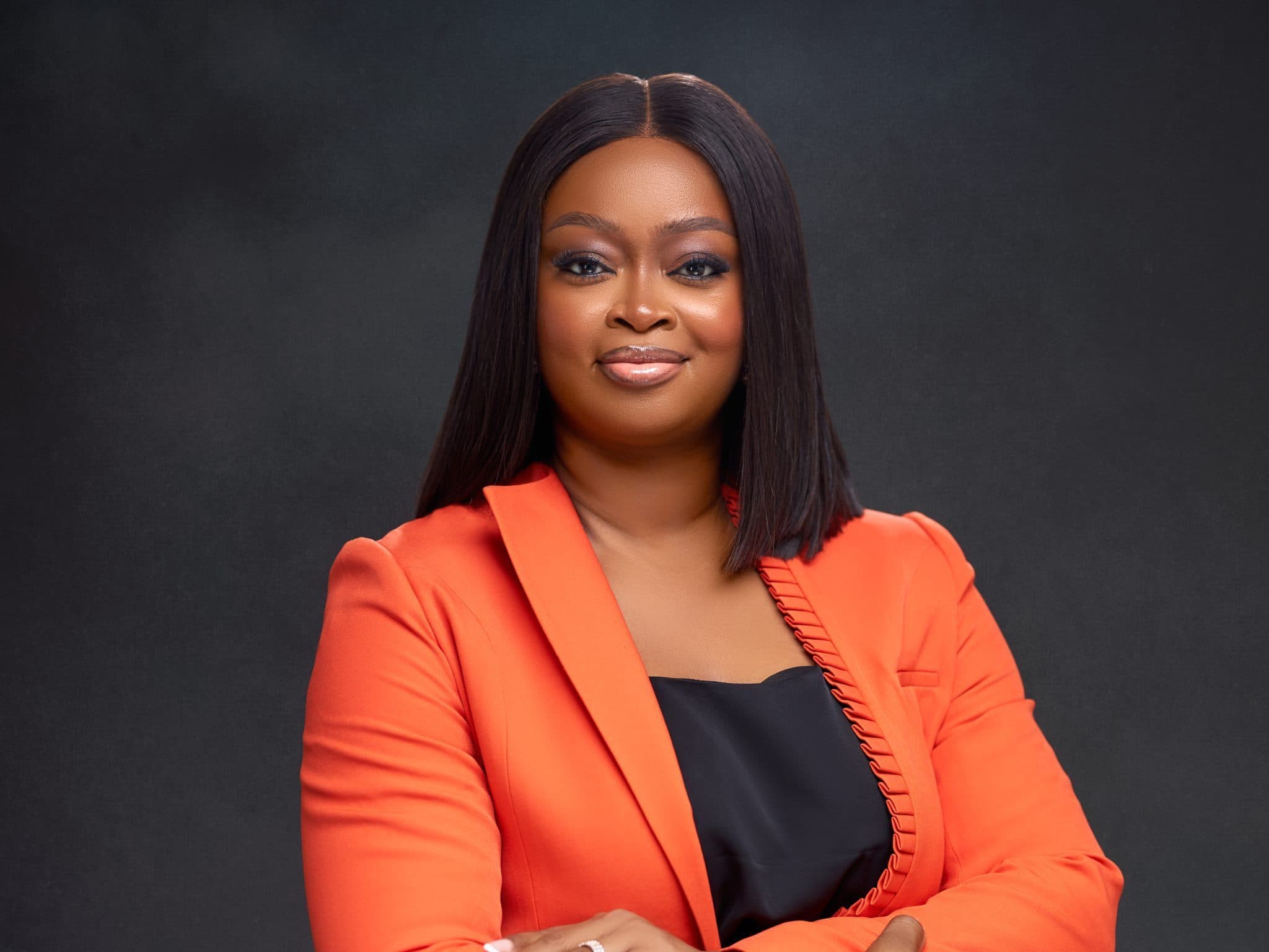 Uche Ajaegbu-bright, arms crossed, in an orange blazer against a dark backdrop